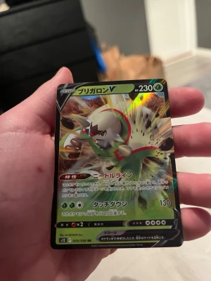 Japansk Pokémonkort Chesnaught V - Säljer ett japanskt Pokémonkort med Chesnaught V-motiv. Kortet har en glansig yta med färger i grönt, vitt och rött samt detaljer i svart. Kortet är tillverkat i laminerad papp och har japansk text. Perfekt för samlare eller Pokémonfans.