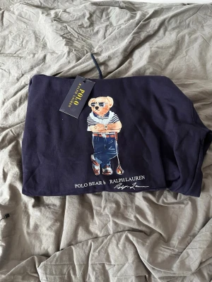 Marinblå hoodie Polo Ralph Lauren - Snygg marinblå hoodie från Polo Ralph Lauren med den ikoniska Polo Bear-trycket framtill. Hoodien har huva med dragsko och är tillverkad i mjukt bomullsmaterial. Perfekt för en avslappnad och trendig stil.