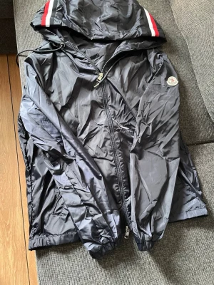 Svart vindjacka från Moncler - Snygg svart vindjacka från Moncler med huva och dragkedja. Jackan har vita och röda ränder på huvan samt Moncler-logga på ärmen. Tillverkad i lätt och glansigt material, perfekt för blåsiga dagar. Två sidofickor med dragkedja och justerbar huva.