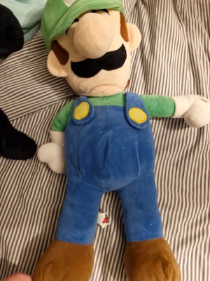 Luigi mjukisdjur med blå hängselbyxor - Stor Luigi-mjukis från Super Mario med grön keps, blå hängselbyxor, grön tröja och bruna skor. Tillverkad i mjukt tyg och har broderade detaljer som mustasch och knappar. Perfekt för alla Nintendo-fans!