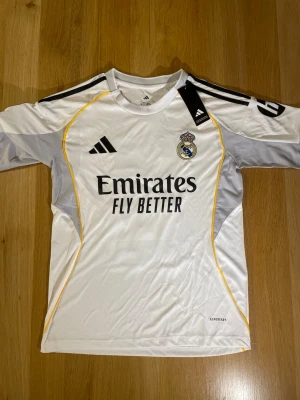 Real Madrid matchtröja Adidas vit - Snygg vit Real Madrid matchtröja från Adidas med klubbmärke på bröstet och svarta detaljer. Tröjan har gula linjer längs sidorna och grå partier på ärmarna. Materialet är lätt och andas, perfekt för fotbollsträning eller match.
