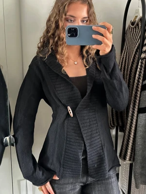 Svart kofta med träknapp - Snygg svart kofta med bred krage och dekorativ träknapp framtill. Koftan har långa ärmar och ribbade detaljer längs kanterna. Perfekt att slänga över en topp för en chill look.mer som M än L