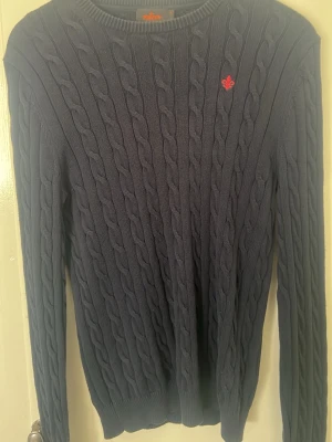 Mörkblå kabelstickad tröja Morris - Mörkblå kabelstickad tröja från Morris med rund halsringning och ribbade muddar. Tröjan har ett rött broderat emblem på bröstet och är tillverkad i mjukt stickat material. Perfekt för dig som gillar klassisk och stilren look.