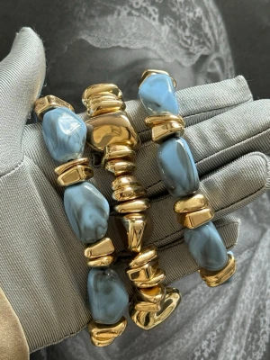 Chunky armband med blå pärlor och guld - Stilrent chunky armband med stora blå marmorerade pärlor och glansiga guldfärgade detaljer. Armbandet har en cool och modern design som sticker ut och passar perfekt för dig som gillar statement-smycken. Materialet är troligtvis plast eller resin och metall.