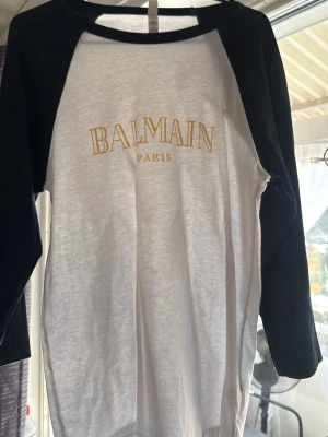 Balmain Paris långärmad t-shirt - Säljer en halvlångarmad t-shirt från Balmain Paris med svart och vit färgkombination. Trycket på bröstet är i guld och ger en exklusiv känsla. Perfekt för dig som vill ha en stilren och trendig look. Materialet känns mjukt och bekvämt. Nypris 3000 kr, den är knappt använd men har inget kvitto kvar därav det billiga priset 
