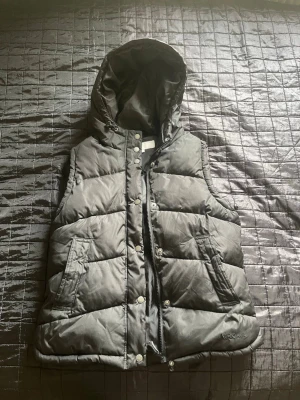 Svart dunväst med huva från northbrooks sports - Snygg svart dunväst från northbrooks med avtagbar huva, dragkedja och tryckknappar framtill. Västen har två snedställda fickor med lock och quiltad design. Perfekt för lager-på-lager och håller dig varm under kyliga dagar. Storlek S/m