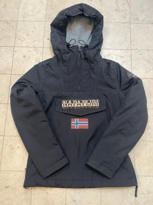Napapijri Jacka - Snygg svart vindjacka från Napapijri med stor huva, halv dragkedja och justerbar dragsko. Insidan är fodrad. Endast använd ett fåtal fall perfekt för våren och lite kyligare dagar.