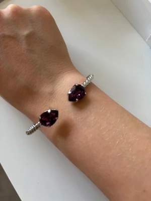 Silverfärgat armband med lila stenar - Snyggt armband i silverfärgad metall med tvinnad design och två stora lila stenar i ändarna. Armbandet är öppet och lätt att justera runt handleden. Perfekt accessoar för att ge din outfit lite extra färg och bling. Armbandet är från caroline svedbom. Pris går att diskutera!🥰