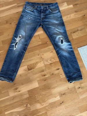 Blå slitna jeans från Nudie Jeans - Säljer ett par blå jeans från Nudie Jeans med tydliga slitningar och hål på båda benen. Jeansen har klassisk femficksmodell, raka ben och är gjorda i denim. Perfekta för dig som gillar en avslappnad och trendig stil.