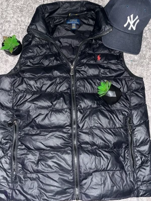 Polo Ralph Laurent väst  - Snygg svart dunväst från Polo Ralph Lauren med hög krage och dragkedja framtill. Västen har två fickor med dragkedja och den klassiska röda loggan broderad på bröstet. Perfekt för lager på lager och streetwear-vibe. 🕺❤️