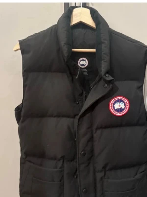 Svart dunväst från Canada Goose - Snygg svart dunväst från Canada Goose med hög krage och klassisk logotyp på bröstet. Västen har tryckknappar, dragkedja och två stora fickor framtill. Perfekt för lager på lager och riktigt varm under kalla dagar.