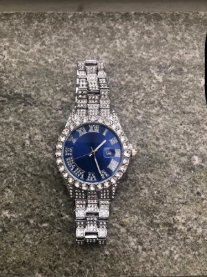 Silverfärgad klocka med blå urtavla - Snygg klocka med glittrande stenar på både armband och boett. Urtavlan är djupblå med silverdetaljer och datumvisning. Klockan har en lyxig känsla och passar perfekt för dig som gillar att sticka ut.