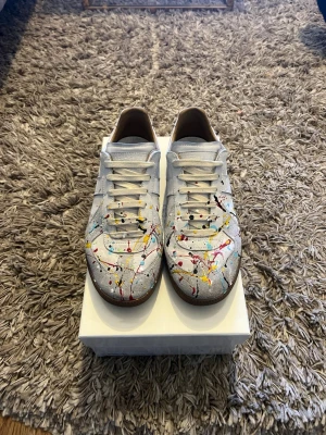 Maison Margiela sneakers med färgstänk - Unika sneakers från Maison Margiela i vitt läder med färgstänk i rött, gult, blått och svart. Klassisk låg modell med gummisula och vita skosnören. Perfekt för dig som vill sticka ut med en kreativ och trendig look. Kvitto finns