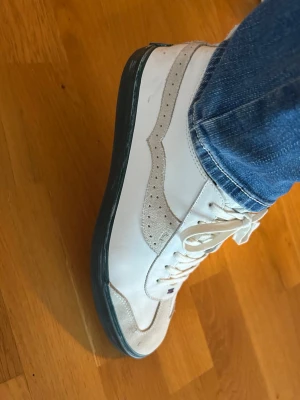 Vita sneakers från Paul Smith  - Snygga vita sneakers från Paul Smith med beige mockadetaljer och klassisk låg siluett. Skorna har vita snören, grön/röd sula och diskret logga på plösen. Perfekta för en clean och stilren look.