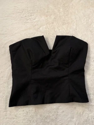 Svart axelbandslös topp - Snygg svart axelbandslös topp med unik v-ringad skärning framtill. Toppen har en croppad passform och är perfekt att styla till både jeans och kjol. Materialet känns stadigt och ger en clean look.