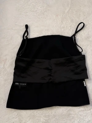 Svart satinlinnetopp från Zara - Snygg svart linnetopp från Zara i satinliknande material. Toppen har smala axelband och en croppad passform med en bred, glansig midjedetalj framtill. Perfekt att styla med jeans eller kjol för en trendig look.