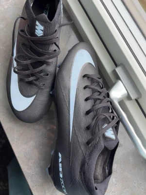 Nike Air Zoom Mercurial fotbollsskor - Snygga Nike Air Zoom Mercurial fotbollsskor i svart med ljusblå detaljer och stor Swoosh-logga. Skorna har snörning, låg profil och greppvänlig yttersula med dobbar för bra fäste på planen. Tillverkade i syntetmaterial för lätt känsla och snabbhet.