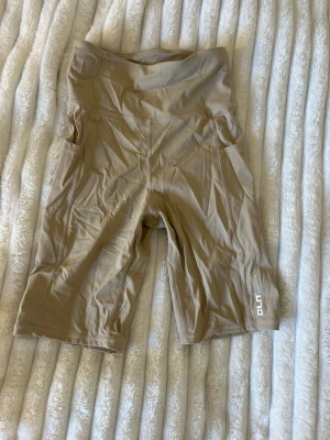 Beige träningstights shorts CLN med fickor - Snygga beige träningstights shorts från CLN Athletics med sidofickor. De är högmidjade och har en tight passform som sitter skönt vid träning. Tillverkade i stretchigt syntetmaterial som andas bra. Perfekta för gymmet eller löprundan. Perfekt för studenten eller alla tillgällen i sommar när man vill ha kjol eller klänning. Helt oanvänd. 