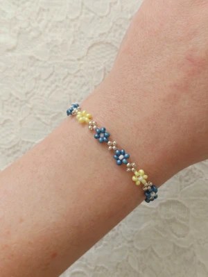 Handgjort blommigt pärlarmband – Blått & Silver – Daisy Chain - handgjort pärlarmband med ett detaljrikt blommönster. En perfekt accessoar som ger en unik touch till både vardag och fest! • Design: Små blommor i en snygg färgkombination av midnattsblått/skimmerblått och ljusgult. Mellan varje blomma sitter dekorativa silverfärgade pärlor. • Detaljer: Silverfärgat klolås med tillhörande förlängningskedja för enkel justering av storleken. • Stil: Klassisk, somrig och tidlös. Matchar utmärkt till jeans eller sommarklänningen. • Skick: Helt nytt/oanvänt