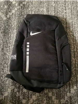 Svart Nike Elite ryggsäck - Snygg svart Nike Elite ryggsäck med vit logga och reflekterande detaljer. Ryggsäcken har flera fack, vadderade axelband med ELITE-tryck och smarta dragkedjor. Perfekt för skolan eller träningen, rymmer mycket och är riktigt bekväm att bära.