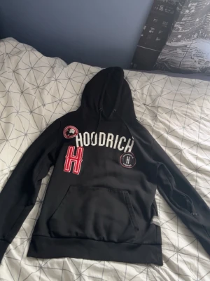 Svart hoodie från Hoodrich med patches - Säljer en svart hoodie från Hoodrich med stora broderade patches och tryck på bröstet. Hoodien har huva med magficka och en extra ficka med dragkedja på ärmen. Perfekt för dig som gillar streetwear och vill sticka ut lite extra.