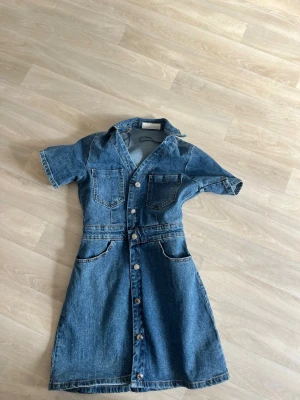 Jeansklänning med knappar från Zara - Säljer en blå jeansklänning från Zara med korta ärmar, krage och knappar hela vägen framtill. Klänningen har två bröstfickor och två större fickor vid höfterna. Perfekt för dig som gillar en avslappnad men snygg look.