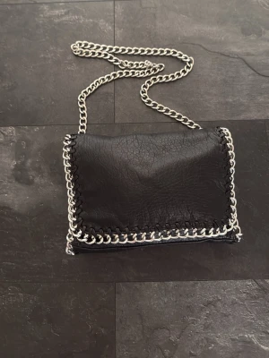 Svart handväska med kedja från Tiamo - Snygg svart handväska från Tiamo i fejkläder med silverfärgad kedja som axelrem och dekorativ kedjedetalj runt kanterna. Väskan har magnetlås och ett innerfack med dragkedja. Perfekt för dig som gillar en edgy och trendig look.
