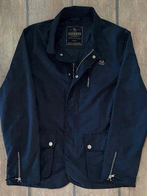 Morris Field Jacket - Riktig snygg och stilren marinblå jacka från Morris med klassisk krage, två stora fickor framtill med tryckknappar, och dragkedjor vid ärmsluten. Jackan har en liten patch med märket på bröstet. Perfekt till våren och kyligare sommardagar. Två innerfickor. Extra knappar finns kvar.