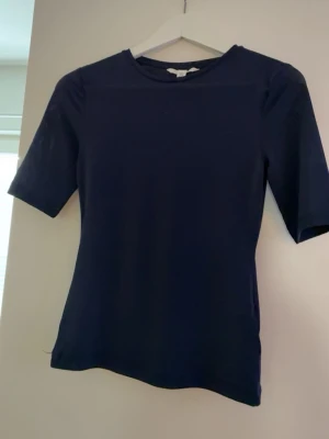 Mörkblå enkel t-shirt från H&M - Snygg mörkblå t-shirt från H&M i enkel och stilren design. T-shirten har korta ärmar och rund halsringning. Tillverkad i mjukt och stretchigt material som känns skönt mot huden. Perfekt basplagg att matcha med allt.