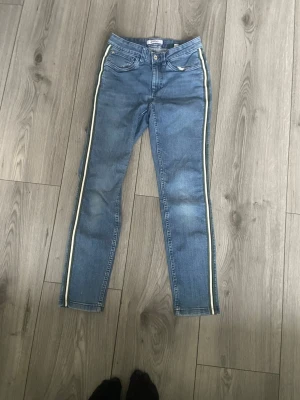 Blå jeans med vita sidoränder - Snygga blå jeans med gröna och vita ränder längs sidorna. Klassisk femficksmodell och normal midja. Jeansen har dragkedjegylf och är tillverkade i ett stretchigt denimtyg för extra komfort.