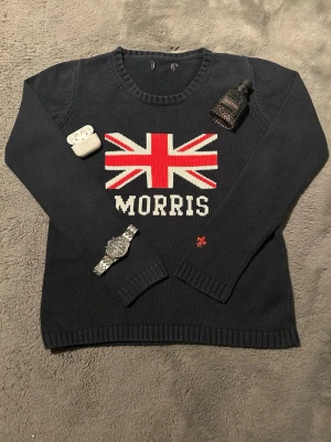 Svart stickad tröja Morris - Svart stickad tröja från Morris med Union Jack-flagga och texten 'MORRIS' framtill. Tröjan har rund halsringning, långa ärmar och ribbade muddar. Materialet är mjukt och passar perfekt för kyligare dagar. Liten röd detalj broderad vid ärmslutet.