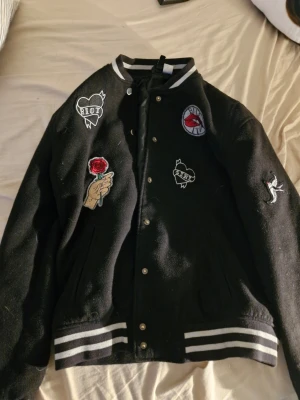 Svart bomberjacka med patches - Svart bomberjacka med vita ränder på muddar och krage. Jackan har flera coola patches, bland annat en röd ros, texten 'RIOT', en hand och ett märke med röda läppar. Stängs med tryckknappar framtill och har en klassisk college-look. Detta är en kvinnlig jacka