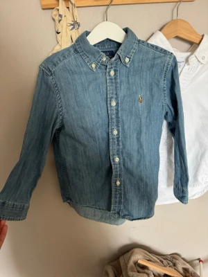 Blå jeansskjorta från Ralph Lauren - Klassisk blå jeansskjorta från Ralph Lauren med button-down krage och broderad logga på bröstet. Skjortan har långa ärmar, knappar framtill och är tillverkad i mjuk bomull. Perfekt för en avslappnad men ändå stilren look.