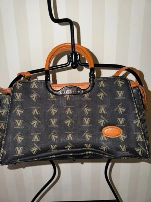 Walter Valentino handväska med logomönster - Snygg handväska från Walter Valentino med svart bas och beige logomönster över hela väskan. Väskan har detaljer i brunt läder, både på handtag och kanter, samt en oval läderpatch med präglad logga. Praktisk axelrem och klassisk, rektangulär form.