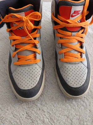 Nike grå och svarta high sneakers - Nike sneakers i high-modell med grå och svarta paneler, orange snörning och detaljer samt perforerad tå. Klassisk Nike-logga på sidan och röd etikett på plösen. Ovandel i läder och syntetmaterial för en cool och sportig look.