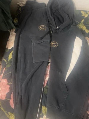 Nike Air mörkblå loungewear set - Mörkblått loungewear set från Nike Air med matchande hoodie och byxor. Hoodien har vit detalj på ärmen och ficka framtill, medan byxorna har sidoficka och vit rand längs benet. Båda delarna har Nike Air-logga i vitt och guld. Perfekt för chill dagar hemma.