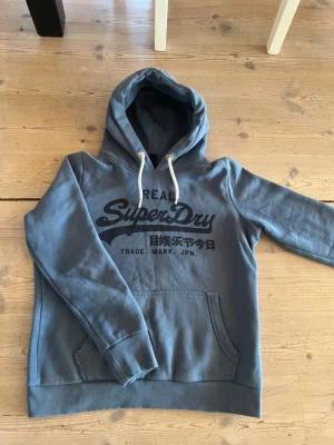 Superdry Hoodie - Säljer en blå hoodie från Superdry med stor logga och japanska tecken på bröstet. Hoodien har vit dragsko, ribbade muddar och en stor magficka. Materialet är mjukt och fleecigt på insidan, perfekt för chill dagar.