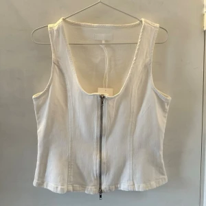 Ny vit denim top  - Säljer en vit denim crop top med djup urringning och dragkedja framtill. Toppen har breda axelband och i en denimlikande material. Den är lite större i storleken. Passar både 38 och 40 beroende på hur man vill den ska sitta. Helt nytt plagg💛