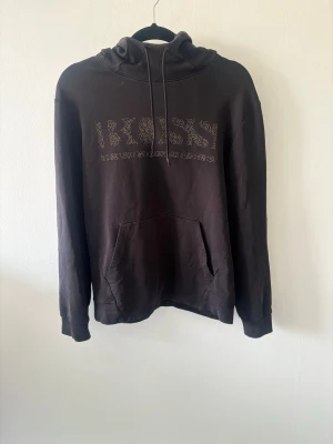 Svart hoodie från BOSS med logga - Snygg svart hoodie från BOSS med stor logga framtill i ett mönstrat tryck. Klassisk känguruficka och justerbar huva med dragsko. Perfekt för en avslappnad och trendig stil. Tillverkad i mjukt bomullsmaterial för extra komfort.