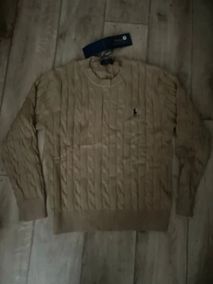 Beige kabelstickad tröja Polo Ralph Lauren - Snygg beige kabelstickad tröja från Polo Ralph Lauren med rund halsringning och diskret logga broderad på bröstet. Tröjan har ribbade muddar vid ärmslut och nederkant. Perfekt för dig som gillar klassisk och stilren look.