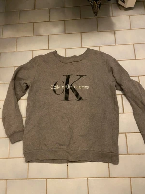 Grå sweatshirt från Calvin Klein - Säljer en grå sweatshirt från Calvin Klein Jeans med stort CK-tryck på bröstet. Tröjan har rund halsringning, långa ärmar och ribbade muddar. Perfekt för en avslappnad och stilren look.