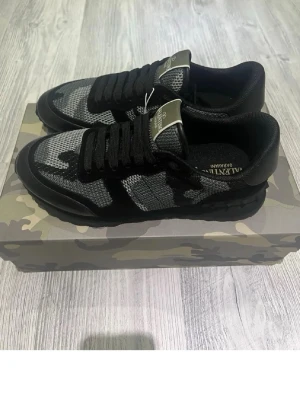 Valentino svarta och grå sneakers - Snygga sneakers från Valentino i svart och grått med kamouflagemönster. Skorna har meshpaneler, svarta läderdetaljer och klassiska svarta snören. Märkeslogga på plösen och innersulan. Perfekta för dig som vill sticka ut med stil.