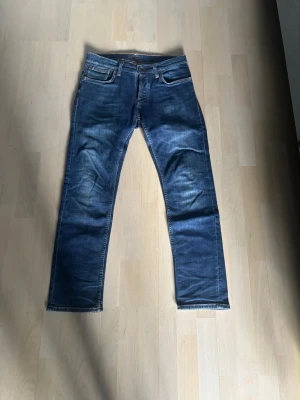 Blå Nudie Jeans - Säljer ett par klassiska blå jeans från Nudie Jeans med raka ben och snygga slitningar. Perfekta för en avslappnad och trendig stil. Storlek: W31 L30