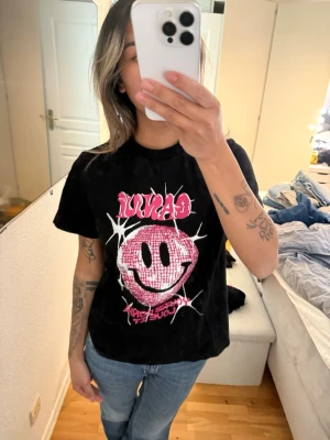 Svart t-shirt med rosa smiley från Ganni - Cool svart t-shirt från Ganni med ett stort rosa smileytryck och text framtill. T-shirten har korta ärmar och en klassisk passform. Perfekt för dig som vill sticka ut med en trendig och lekfull look.