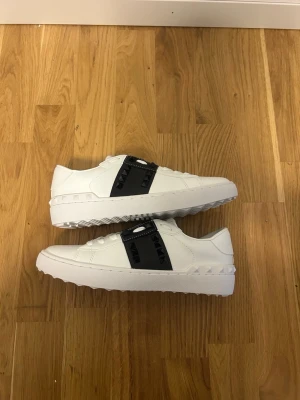Valentino Rockstud Untitled sneakers - Snygga vita sneakers från Valentino med svart band över vristen dekorerat med svarta nitar. Klassisk låg modell med snörning och chunky sula. Skorna är tillverkade i skinn och har en unik, edgy design som sticker ut.
