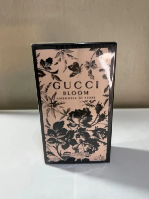 Gucci Bloom Ambrosia di Fiori parfym - Gucci Bloom Ambrosia di Fiori är en lyxig parfym i en elegant rektangulär förpackning med blommigt svart mönster på ljusrosa botten. Flaskan rymmer 100 ml och har en exklusiv känsla, perfekt för dig som gillar unika och sofistikerade dofter.
