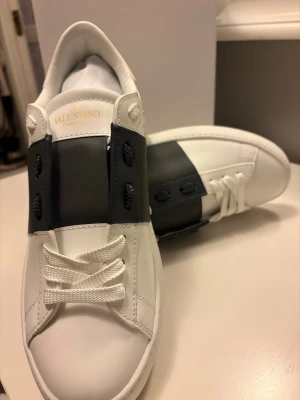 Valentino vita sneakers med mörkblå band  - Snygga vita sneakers från Valentino med ett brett mörkblå band över snörningen. Skorna har klassisk låg profil, vita skosnören och diskret Valentino-logga på plösen. Tillverkade i slätt läder för en clean och modern look.