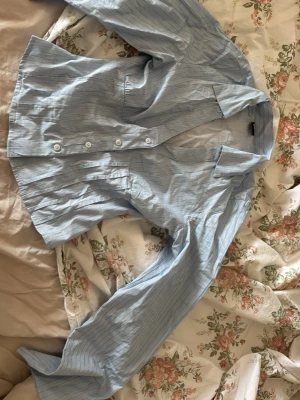 Blårandig skjorta från Gina Tricot - Säljer en ljusblå skjorta från Gina Tricot med tunna vita ränder. Skjortan har klassisk krage, knappar framtill och långa ärmar. Perfekt till jeans eller kjol för en fräsch look.