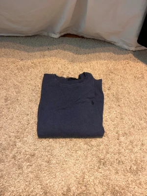 Mörkblå stickad tröja från Polo Ralph Lauren - Mörkblå stickad tröja från Polo Ralph Lauren med rund halsringning och diskret broderad logga på bröstet. Tröjan har långa ärmar och är tillverkad i ett mjukt material som passar perfekt till lager-på-lager-stil. Storlek M