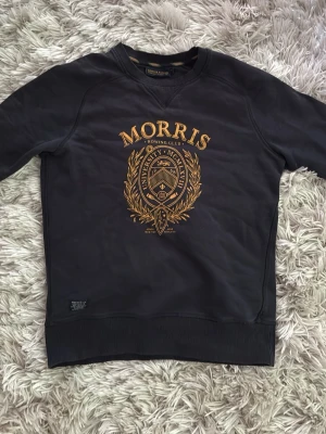 Svart sweatshirt från Morris R. Club - urtvättad lapp. Cool svart sweatshirt från Morris R. Club med stort guldfärgat tryck och emblem på bröstet. Tröjan har rund halsringning och ribbade muddar vid ärmslut och nederkant. Perfekt för en avslappnad och stilren look.